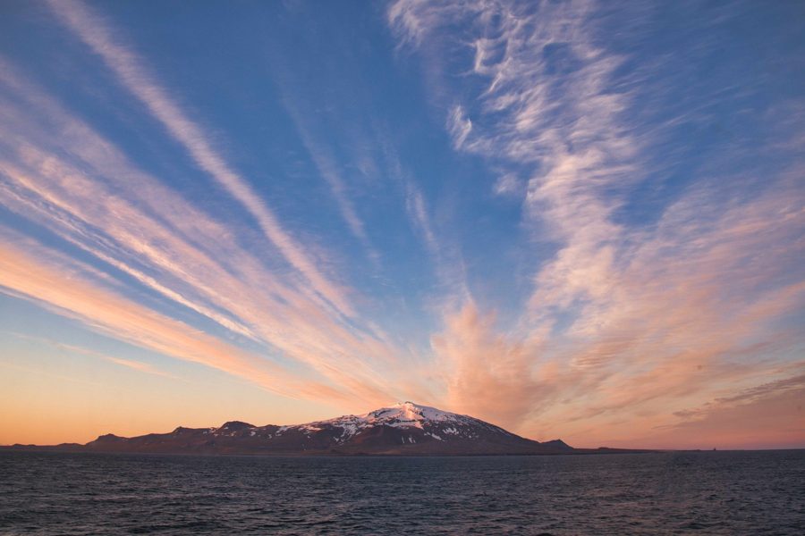 Streaks of cloud glowing pink and orange at sunset over a snow-capped mountain and dark sea